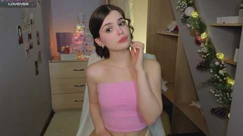 Video of a young woman with light skin and short dark hair, wearing a pink strapless top, sitting in a cozy room with festive decorations, including a Christmas tree.