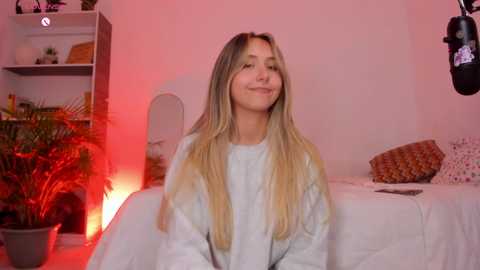 Media: Video of a blonde Caucasian woman in a white robe, smiling, in a cozy bedroom with red lighting, potted plants, and a microphone.