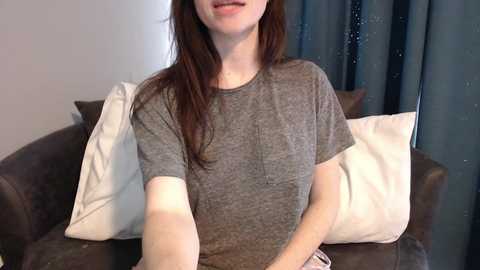 Media: Video of a young woman with fair skin, long brown hair, and a gray t-shirt, sitting on a dark brown leather couch with white pillows, in a dimly lit room.