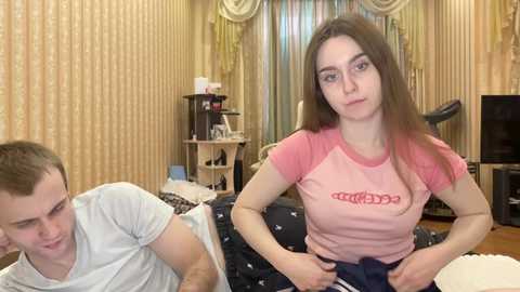 Media: Video of a young, slender, light-skinned woman with brown hair wearing a pink t-shirt, and a young man with short hair in a grey t-shirt. They're in a cluttered, beige-patterned room with a television and a desk.