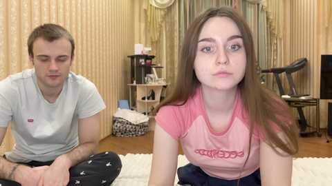 Media: Video of a young Caucasian couple in a cozy bedroom. The woman, with long brown hair and a pink shirt, pouts. The man, with short hair and a gray shirt, sits nearby.