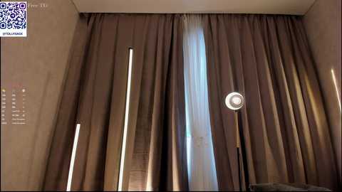 Media: Video of a modern bedroom with brown, floor-length curtains, a vertical light bar on the left, and a circular light on the right. The background wall is beige with a small calendar and QR code in the top left corner.