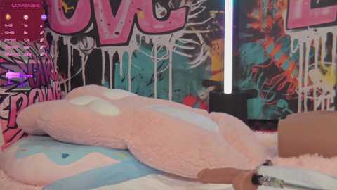 Media: Video of a plush bunny laying on a soft bed in a room with graffiti-covered walls and vibrant decor. The bunny's fur is fluffy and white, with a pink nose and ears.