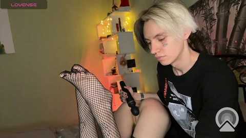 Media: A video of a pale-skinned, androgynous person with platinum blonde hair, wearing a black T-shirt, fishnet stockings, and a harness, using a vibrator in a dimly lit room with a shelf of books and string lights.