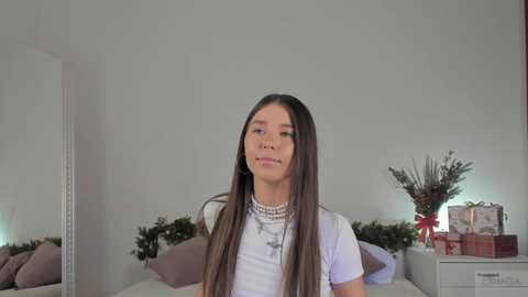 Media: Video of a young woman with long, straight, dark hair, wearing a white blouse and pearl necklace, standing in a minimalistic bedroom with a bed, a Christmas wreath, and a gift box.