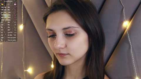 Video of a young woman with fair skin and long dark hair, wearing a sleeveless top, with a serene expression, surrounded by soft yellow fairy lights and a star chart on a dark wall.