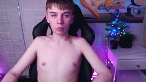 Media: Video of a young, shirtless, Caucasian male with light skin and short brown hair, seated in a black gaming chair. Background features a white tiled wall, a small Christmas tree with blue lights, and a potted plant.