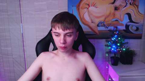 Media: Video of a young, pale-skinned, shirtless boy with short brown hair in a black gaming chair, surrounded by festive lights and a small Christmas tree, against a brick wall and colorful abstract art.