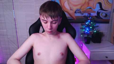 Media: Video of a shirtless, pale-skinned, young boy with short brown hair sitting in a black gaming chair. He has a neutral expression. Background features a white tiled wall, a colorful mural, and a small Christmas tree with multi-colored lights.