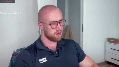 Video of a bald, bearded, fair-skinned man with glasses, wearing a dark polo shirt, sitting indoors.