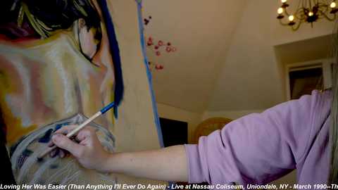 Media: Video of an artist painting a detailed, realistic portrait of a woman's face in a home studio. The artist, wearing a pink shirt, uses a paintbrush on a canvas. The room has a chandelier and a wooden door.