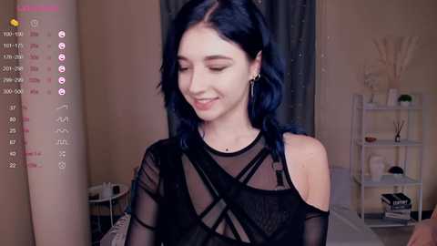 Media: Video of a young woman with dark blue hair, fair skin, wearing a sheer black top, smiling softly in a dimly lit room with white shelves and a plant.