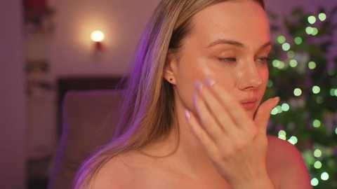 Media: Video of a blonde woman with light skin and closed eyes, touching her face, with a blurred Christmas tree and warm, purple-hued lighting in the background.