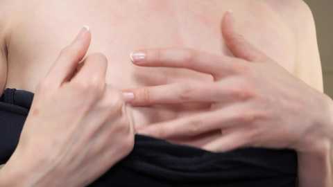 Media: A close-up video of a person's upper body, focusing on the chest. The person is pulling down a black garment to reveal pale, smooth skin. The hands, with light pink nails, are gently touching the skin.