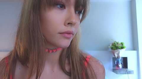 Media: Video of a young woman with long, light brown hair and fair skin, wearing a pink floral top, looking thoughtful. Background features a white shelf with a small plant.