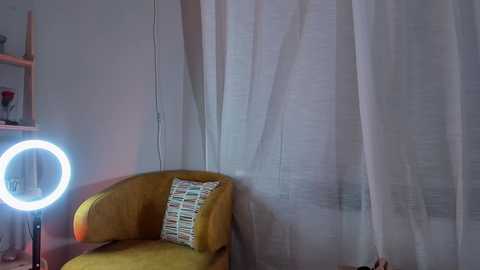 Media: Video of a mustard-yellow armchair with a patterned cushion, white sheer curtains, and a glowing round light bulb on a wooden table in a softly lit room.