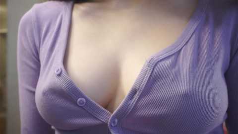 Media: Video of a light-skinned woman with large breasts, wearing a lavender ribbed cardigan with a deep V-neck, revealing cleavage. The background is blurred, suggesting an indoor setting.