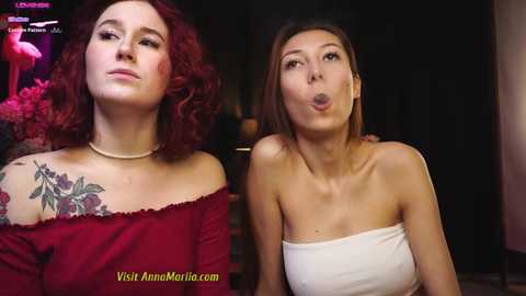 Media: Video of two women: one with curly red hair, off-shoulder red top, floral tattoo, and the other with straight brown hair, white strapless top, puckering lips. Background shows a dimly lit room.