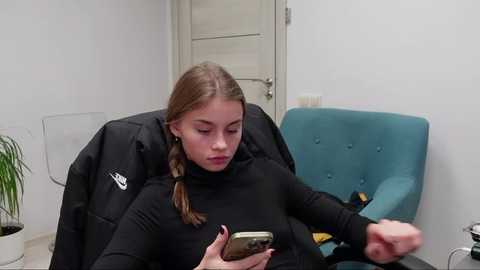 Media: A young woman with light brown hair in braids, wearing a black turtleneck, sits on a teal chair, engrossed in her smartphone. Background features a beige door and a potted plant.