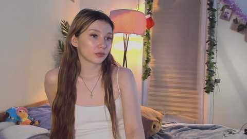 Media: A video of a young Asian woman with long brown hair, wearing a white tank top, sitting on a bed in a dimly lit room with a lamp and a stuffed toy.