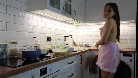 Media: A video of a young Asian woman with long dark hair, wearing a pink apron, standing in a modern white-tiled kitchen with wooden countertops, cooking appliances, and utensils.