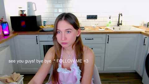 Media: Video of a young woman with long brown hair in a white apron, sitting in a modern kitchen with white cabinets and wooden countertops.