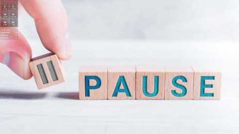 Media: A video featuring a hand holding a wooden block with the word \"PAUSE\" in blue letters. The background is a soft-focus, light-colored, textured wall. The image emphasizes the concept of pausing.