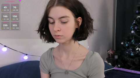 Video of a young woman with shoulder-length dark hair, wearing a grey top, sitting indoors near a Christmas tree and string lights.