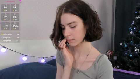 Media: A video of a young woman with short, dark hair, wearing a green top, looking down at her phone, in a dimly lit room with a Christmas tree in the background.