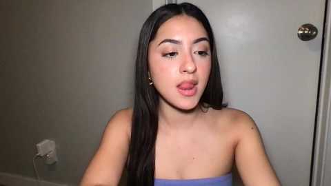 Media: Video of a young Latina woman with long, straight black hair, light skin, and full lips, wearing a strapless blue top, sitting indoors with a plain grey door and white outlet in the background.