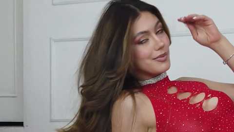 Media: Video of a smiling woman with long, wavy brown hair, wearing a red, sequined, cut-out dress, standing against a white door.