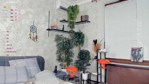 Media: Video of a modern, minimalist bedroom with a grey bed, white walls, green potted plants, a dreamcatcher, and red desk lamps, featuring a calendar and a window with blinds.