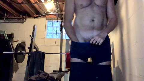 Media: A video shows a shirtless man with fair skin and a lean build, wearing only navy blue briefs, standing in a dimly lit basement with white walls, exposed pipes, and a laundry rack.