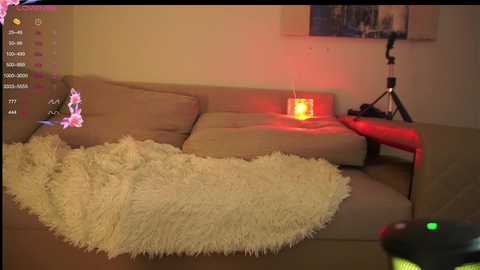 Media: Video of a cozy living room with a beige sectional sofa, a fluffy white throw, a glowing red candle, and a video game controller in the foreground.