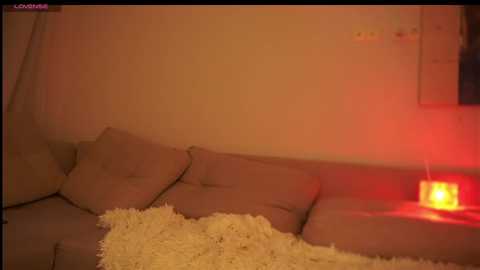 A video of a cozy room bathed in warm, orange light. A beige sofa with cushions and a fluffy, cream-colored blanket. A lit red candle on the right.