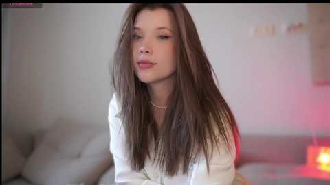 Media: Video of a young, fair-skinned woman with long, straight brown hair, wearing a white blouse and a delicate necklace. She is in a softly lit room with a neutral background and a subtle red glow.