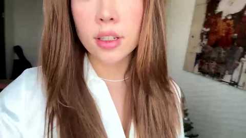 Video of a light-skinned woman with long, straight brown hair, wearing a white blouse and a silver necklace. She has full lips and neutral expression. Background features abstract art with earthy tones.