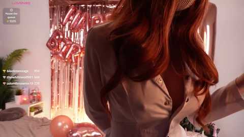 Media: Video of a woman with long, wavy red hair, wearing a beige blazer, standing near a bed with a white sheet and a red balloon. A silver tinsel backdrop with a \"2020\" sign is in the background.