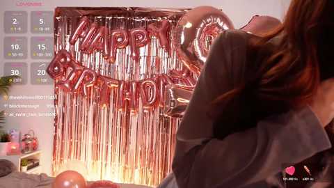 Media: Video of a birthday party with a \"Happy Birthday\" banner and balloons, featuring a woman with wavy brown hair in a gray shirt, background showing a calendar and \"Live\" text.