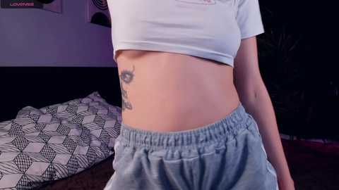 Media: Video of a slender, light-skinned woman with a visible tattoo on her ribcage, wearing a cropped white top and loose, light blue sweatpants, standing in a dimly lit bedroom with patterned bedding and dark furniture.