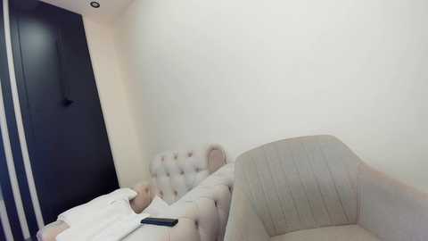 Video of a modern, minimalist hotel room with a beige tufted headboard, a light gray upholstered armchair, and a white pillow. A person is lying in bed with a white blanket and a black remote control on the bedside table.