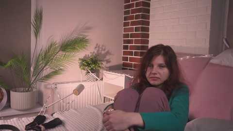 Media: Video of a woman with shoulder-length brown hair, wearing a green top and gray pants, sitting on a white wicker chair, surrounded by plants and a brick wall, reading a book.