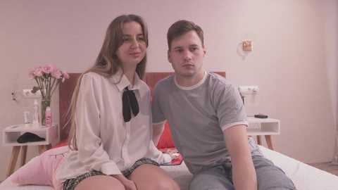 Media: Video of a young Caucasian couple, a brunette woman in a white blouse and plaid skirt, and a man in a gray T-shirt, sitting on a bed with red pillows and a white duvet.