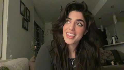 Media: Video of a smiling woman with long, wavy dark hair, wearing a black top, in a modern living room with beige walls and framed photos.