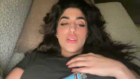 Media: Video of a woman with long, wavy dark hair, light skin, and full lips, lying on a beige couch, wearing a black t-shirt, with her hand on her chest.