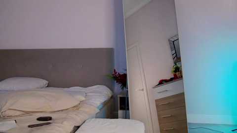 Media: A video of a minimalist bedroom with a beige upholstered headboard, white bedding, a wooden dresser, and a large mirror reflecting a corner. A blue light source illuminates the room.