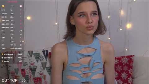 Media: Video of a young Caucasian woman with shoulder-length brown hair, wearing a revealing blue bodysuit with large cutouts, sitting on a sofa with festive decorations in the background.