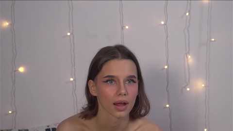 Media: A video of a young woman with fair skin, brown hair, and blue eyes, gazing thoughtfully. She is topless against a backdrop of softly glowing, string fairy lights.