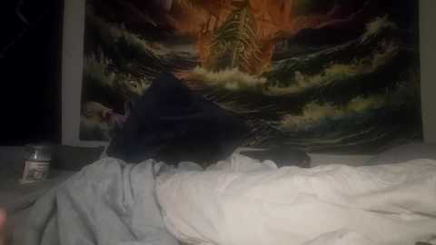 Media: A video of a person in a dark room with a large, colorful, abstract wall tapestry featuring a fiery orange and green pattern. The person is lying in bed, covered by white sheets, with a dark pillow.