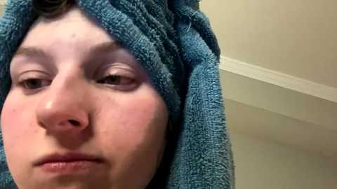 Media: A close-up video of a young boy with fair skin and short brown hair wrapped in a blue towel, showing a neutral expression. The background is a plain, off-white wall with a white ceiling molding.